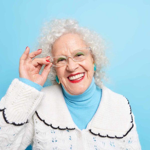 Portrait of good looking cheerful grey haired grandmother smiles toothily keeps hand on rim of spectacles has well cared complexion wrinkled sin dressed in white jumper isolated over blue background